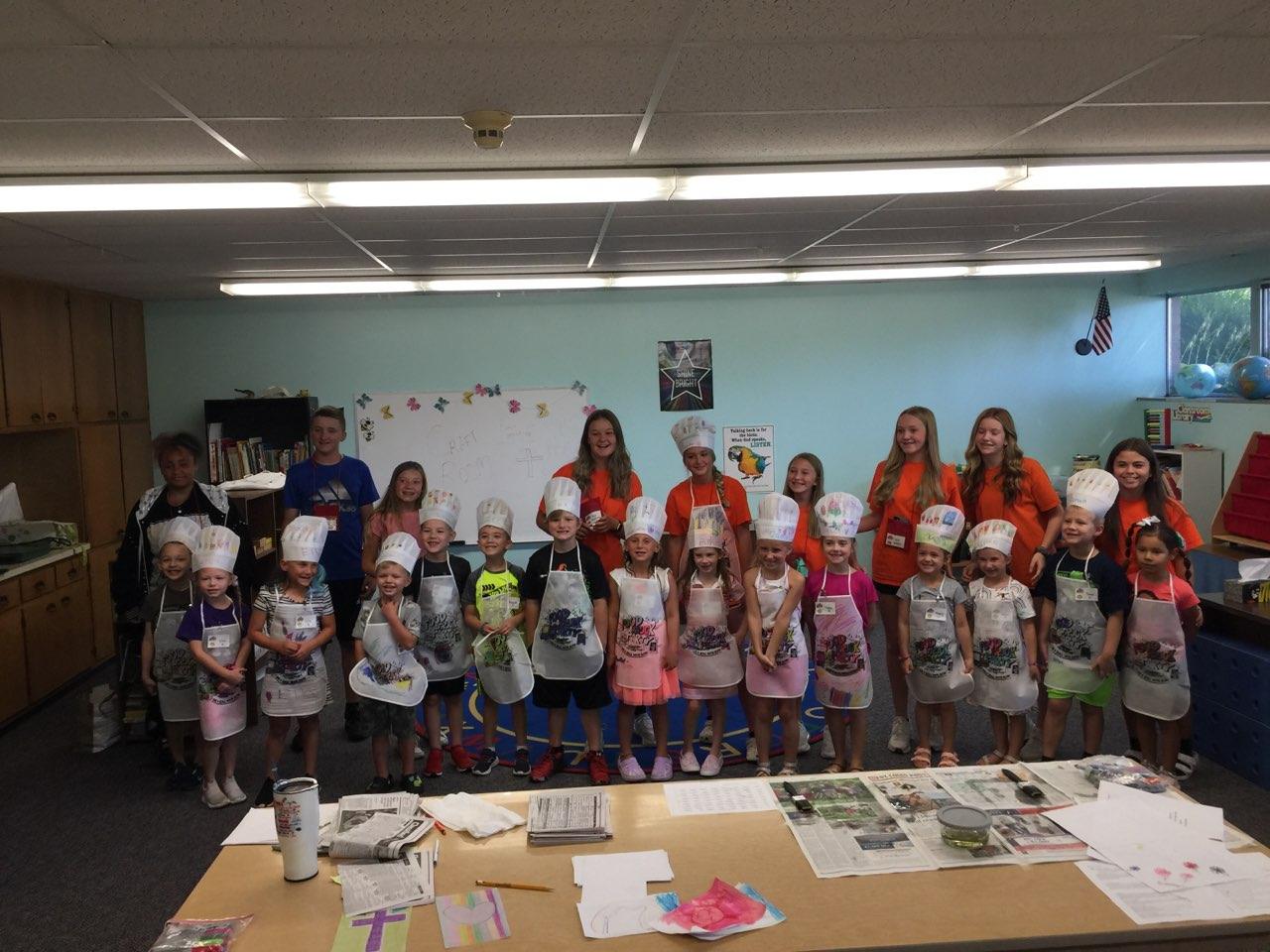 Children and adults in chef hats and aprons in a classroom. Orange shirts and white aprons.