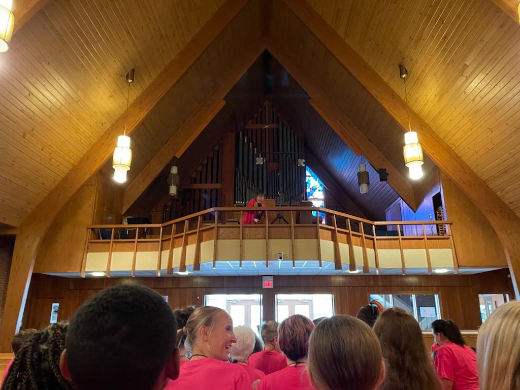 People in pink shirts facing a balcony with a person, inside a church with wooden ceiling.