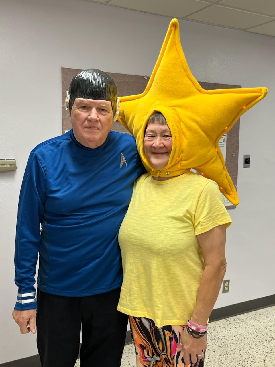Man in Star Trek uniform and woman in a yellow star costume smiling, indoors.