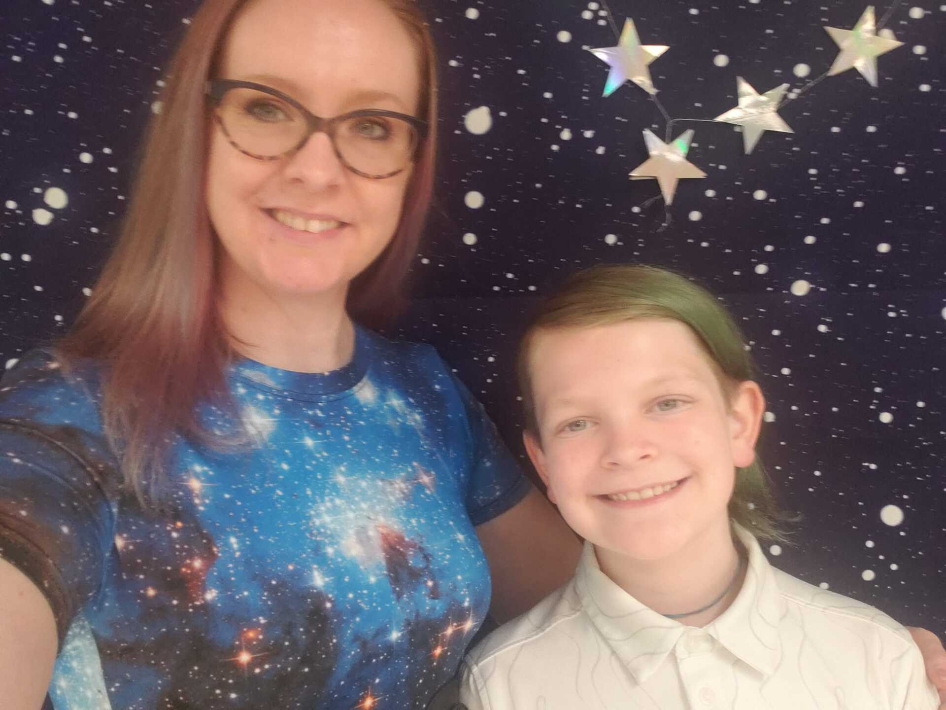 Woman and child smiling, standing in front of a starry background. Woman wears glasses and space shirt; child has green hair.