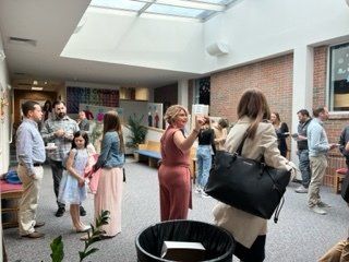People mingling in a bright, open hallway; a woman in pink gestures while others chat.