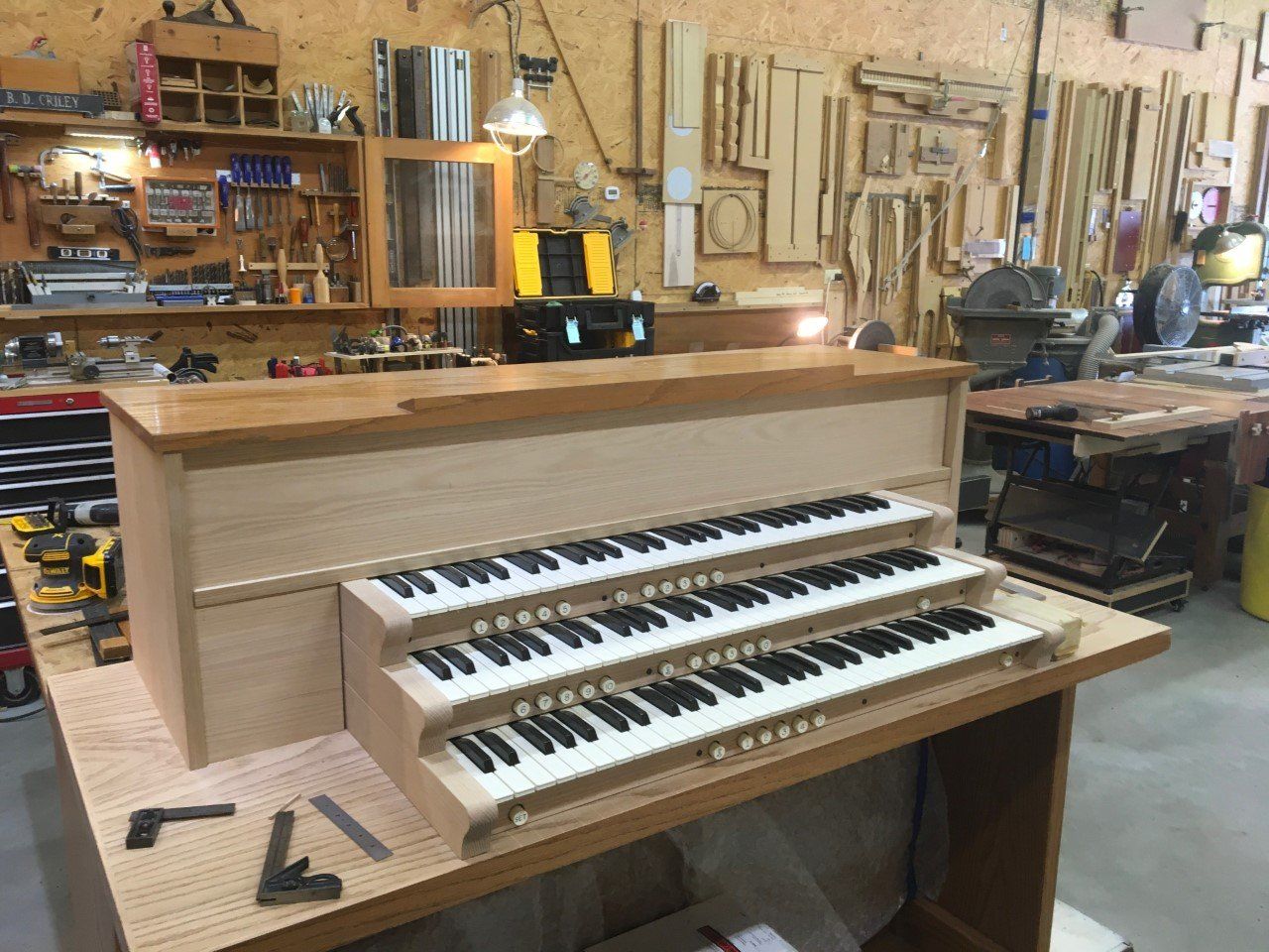 A three-keyboard organ in a wood shop, with tools and work benches in the background.
