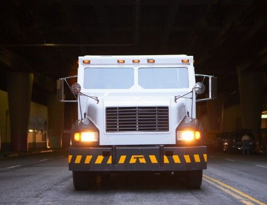 White armored truck driving toward the camera under an overpass with yellow hazard stripes on the front.