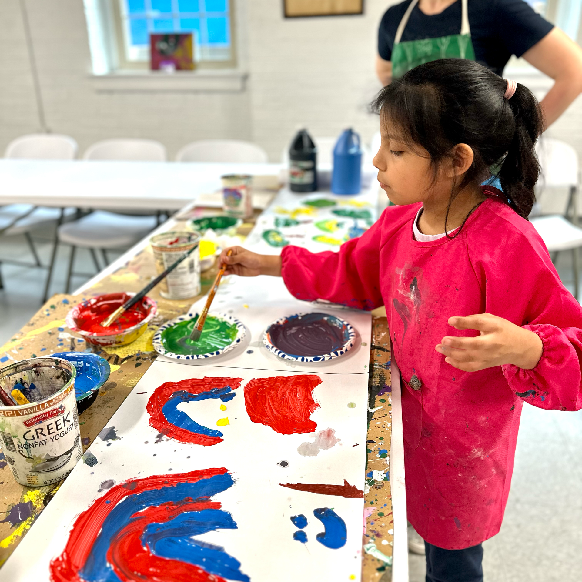 A little girl in a pink apron is painting on a long piece of paper