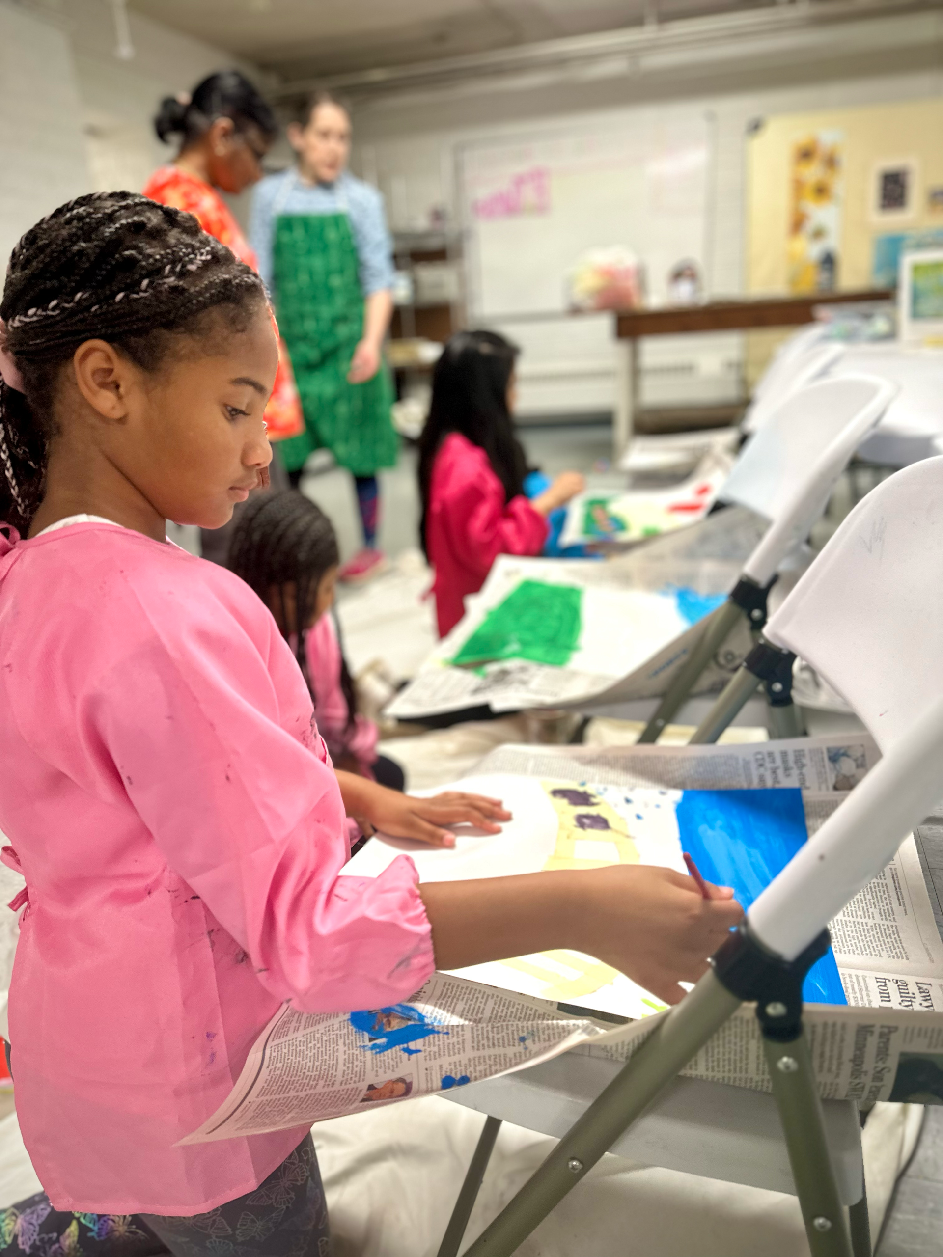 A girl in a pink apron is sitting in a chair painting a picture.