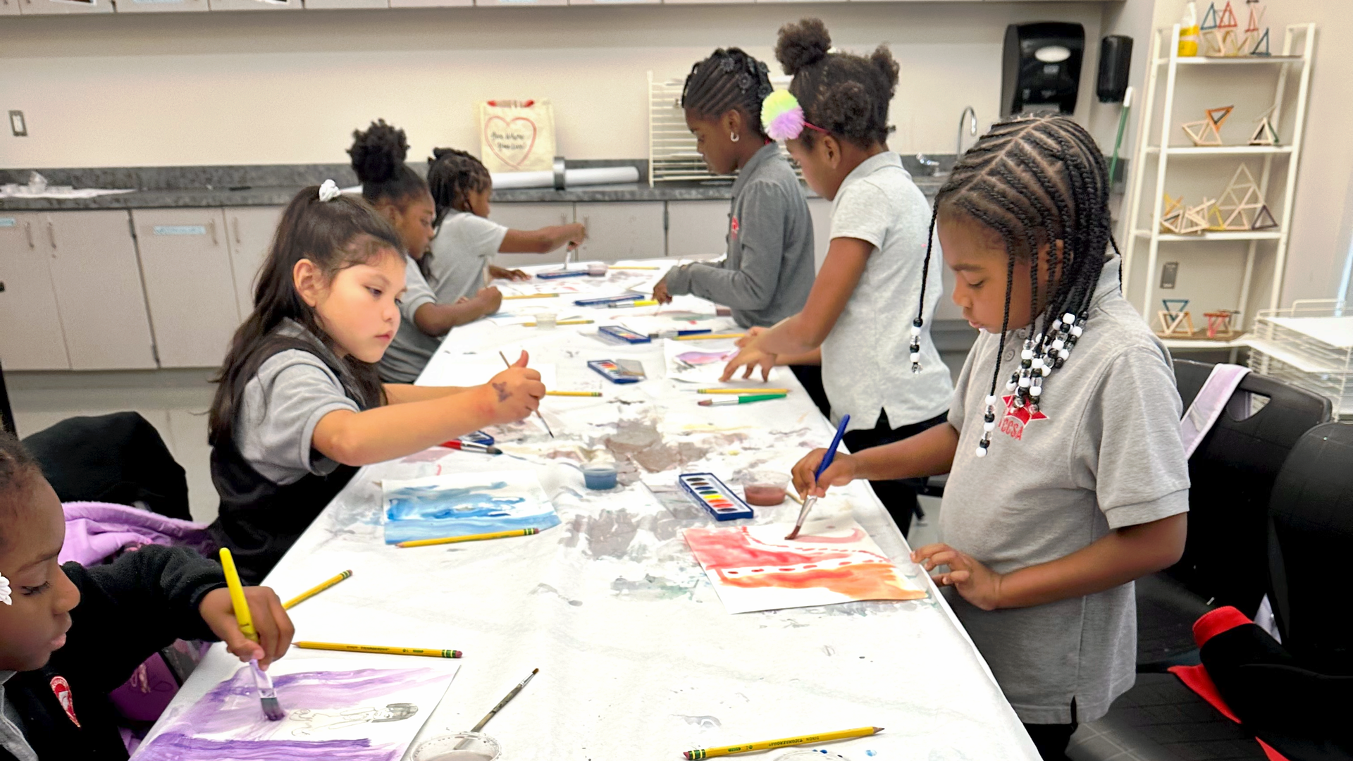 A group of children are sitting at a long table painting.