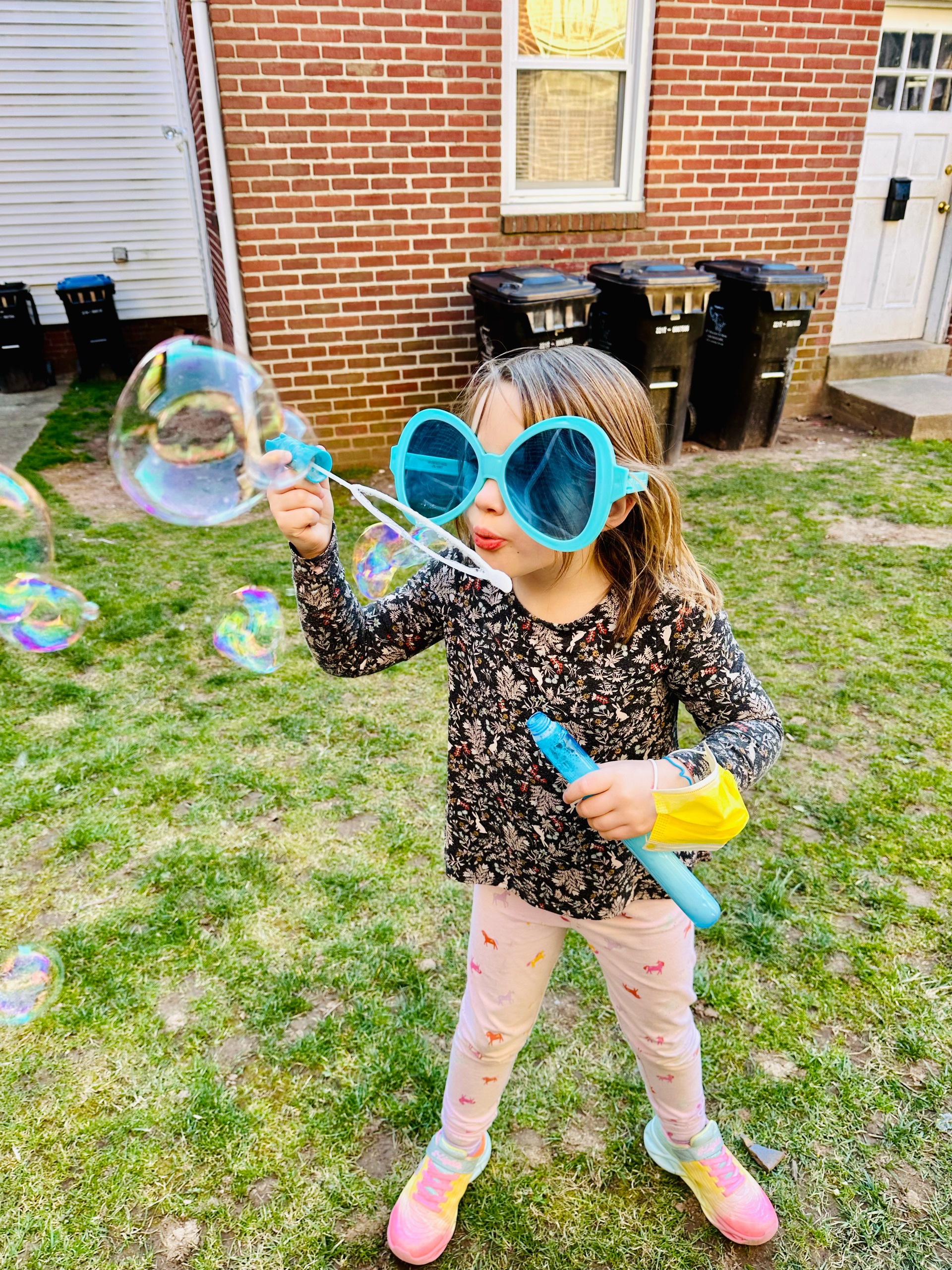 A little girl wearing sunglasses is blowing soap bubbles in a yard.