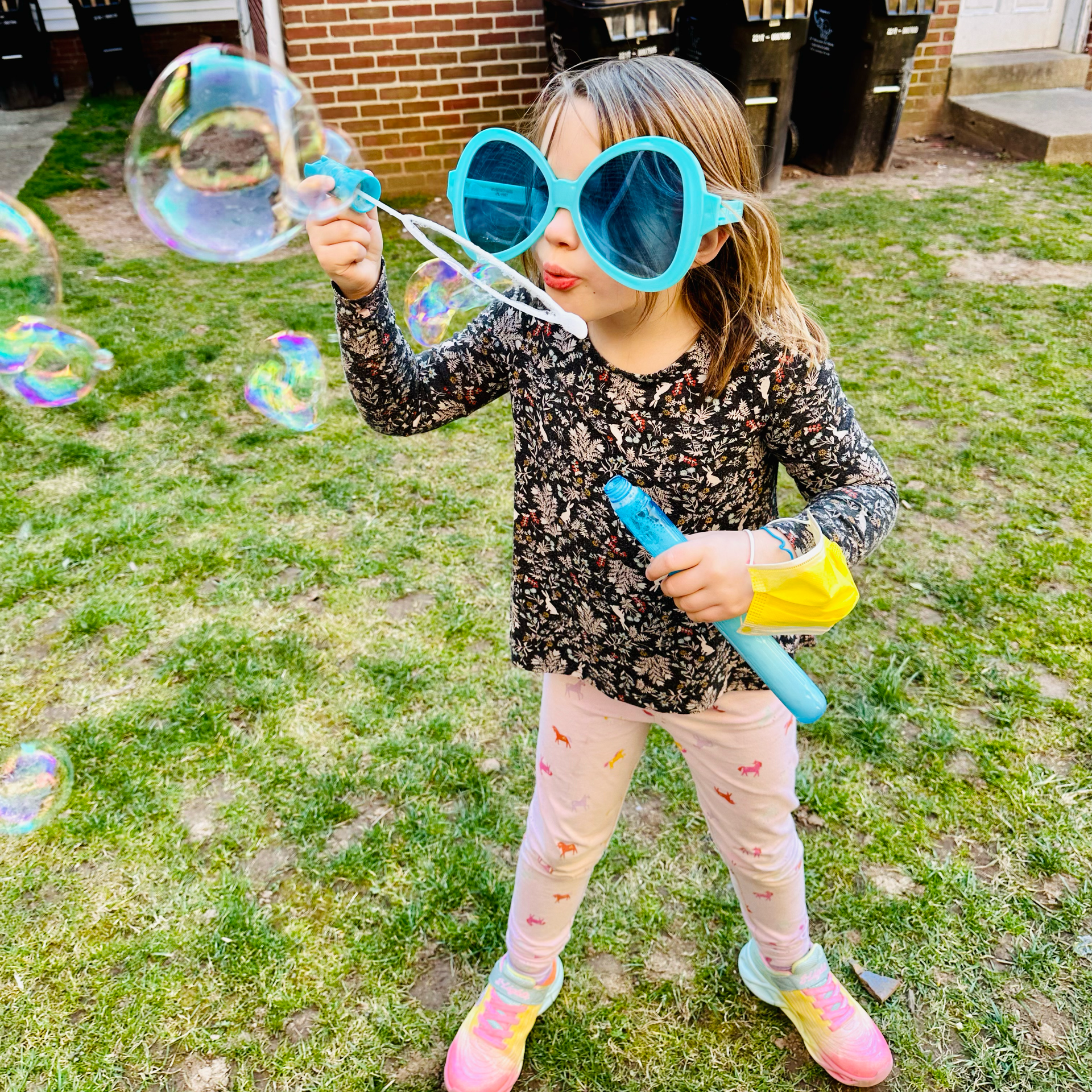 A little girl wearing sunglasses is blowing soap bubbles in a yard.