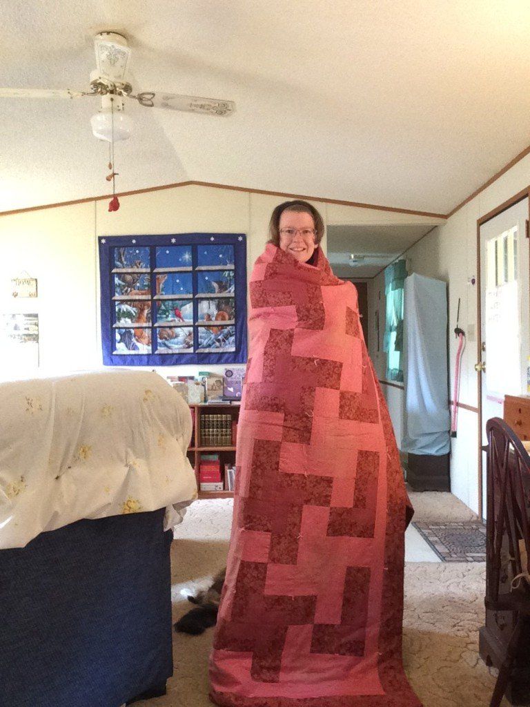 Person wrapped in a pink and burgundy quilt standing in a well-lit living room.