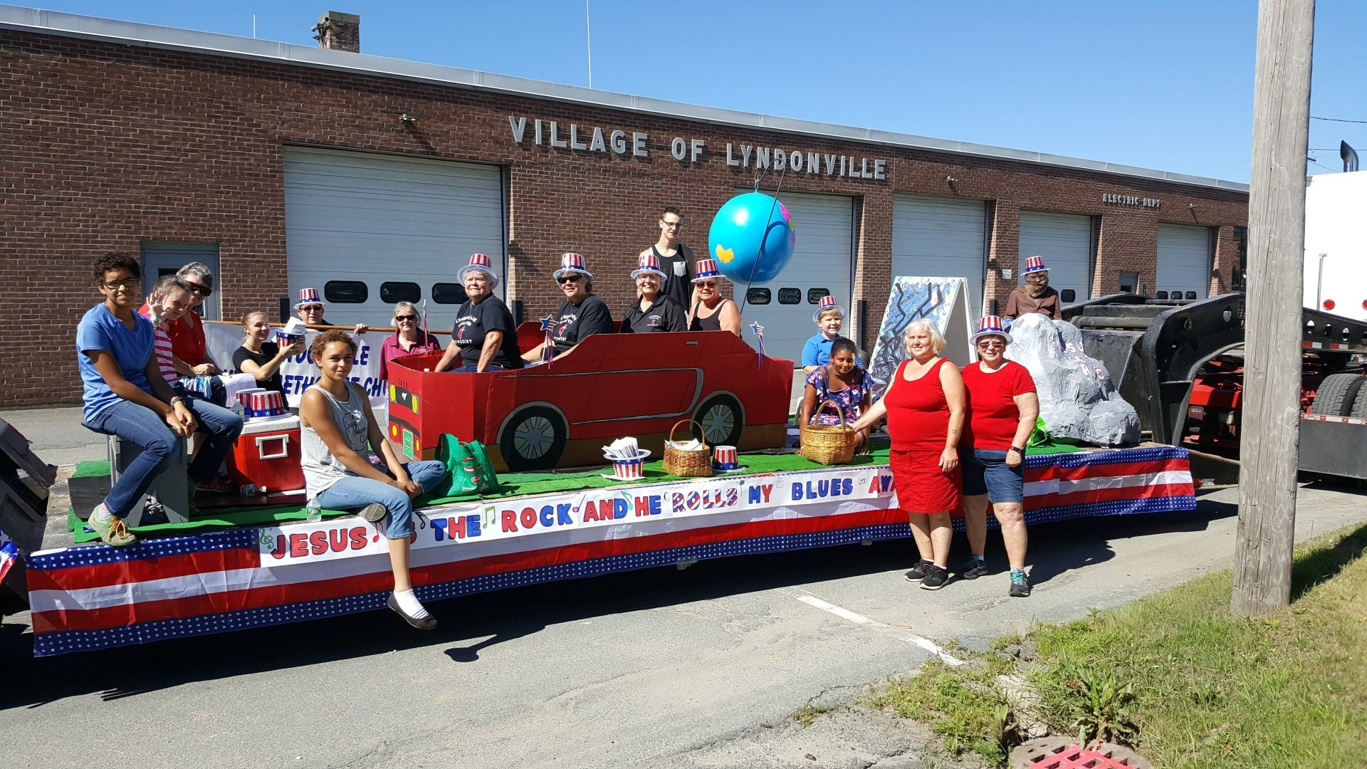 Parade float with people, red car, blue, white, and red decorations in front of a brick building.