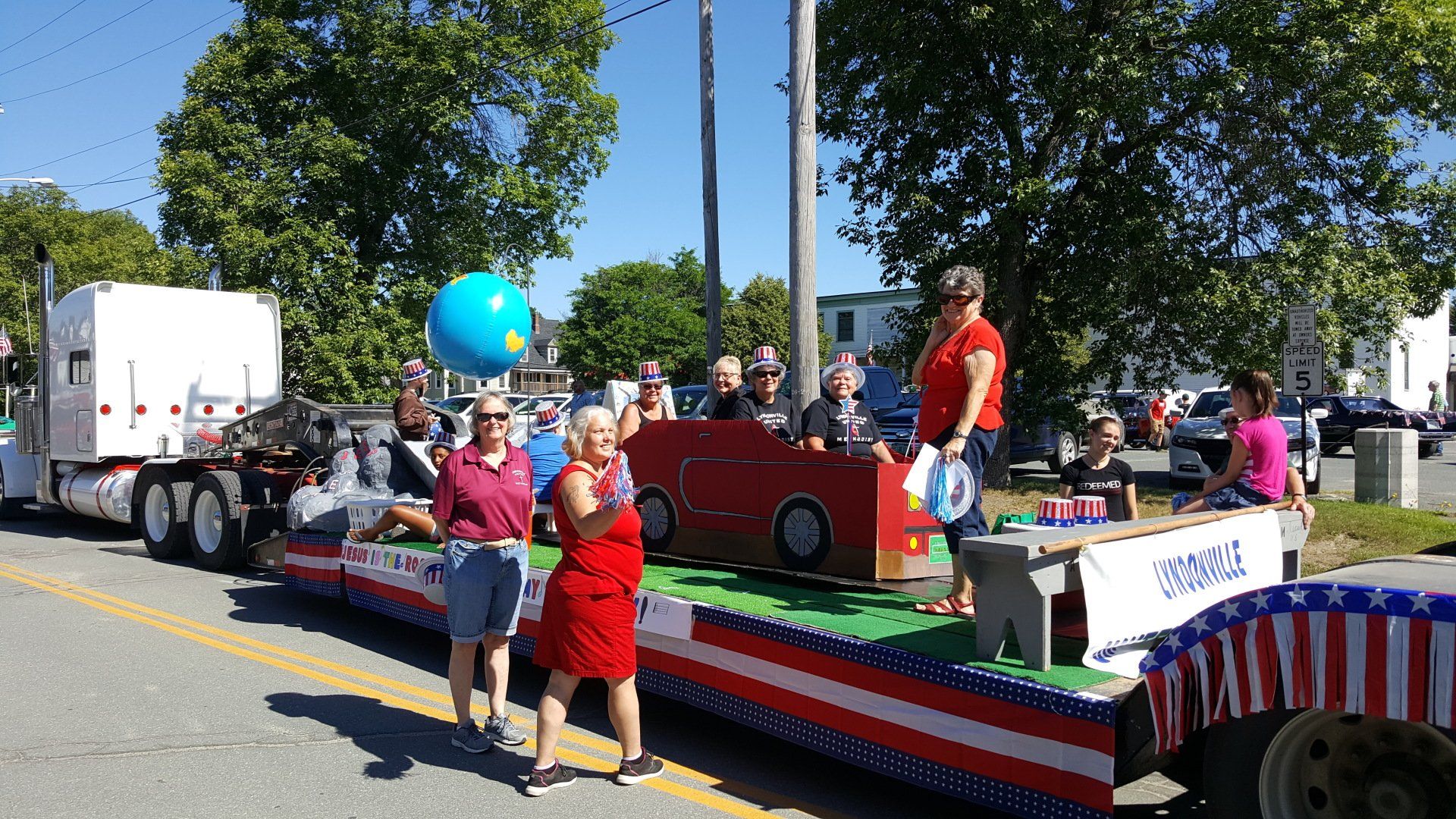 Parade float with people decorated in red, white, and blue, with a cardboard car, on a sunny day.