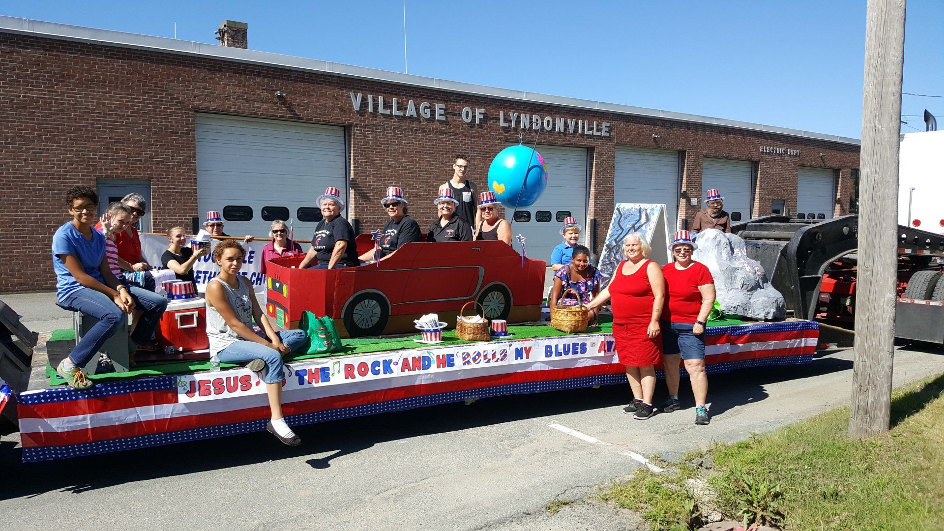 A parade float in front of a brick building. People ride on a red car float with a blue globe.