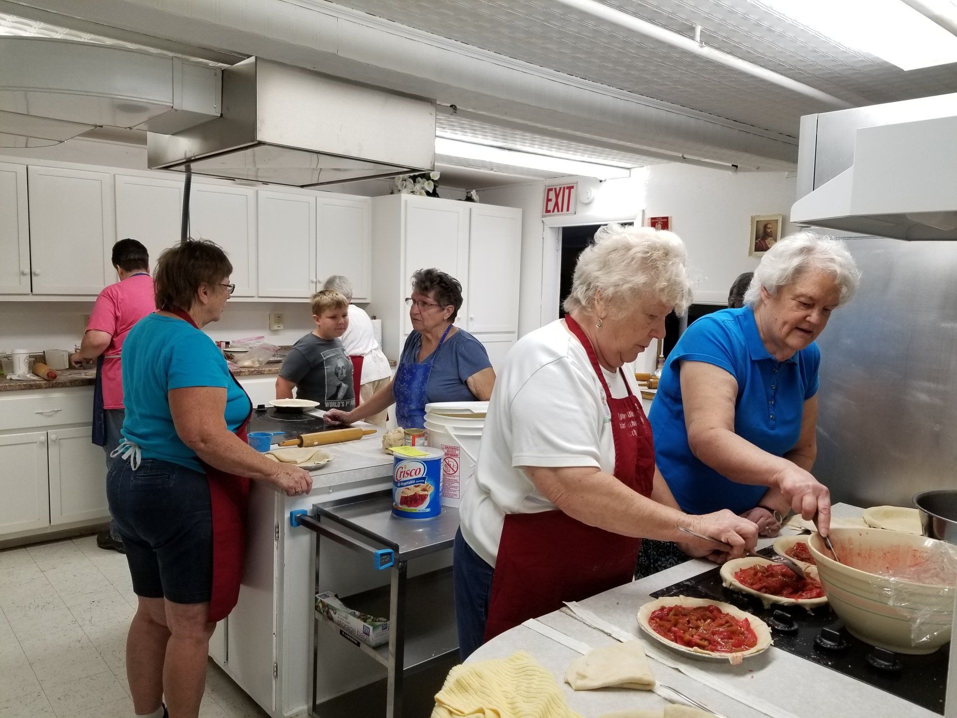 People preparing food in a kitchen. Women wear aprons, work at a counter with pies, and fill ingredients.
