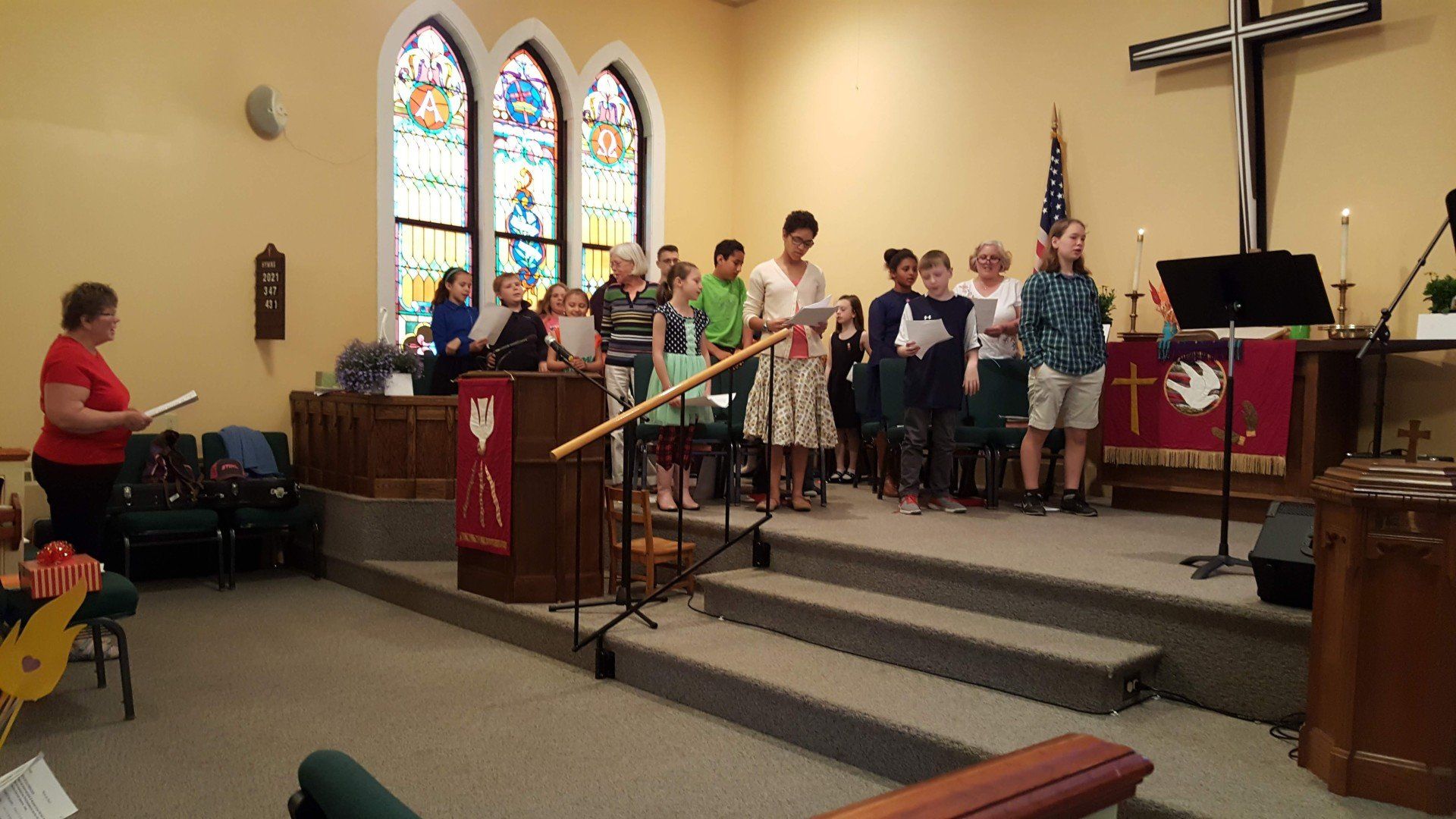 Choir singing at the front of a church, standing on steps, with stained glass windows in the background.
