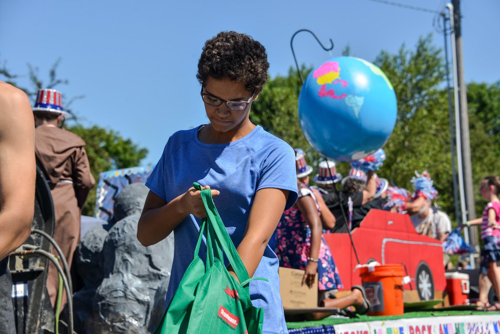 Woman in blue shirt looks into green tote bag near parade float with globe and people.