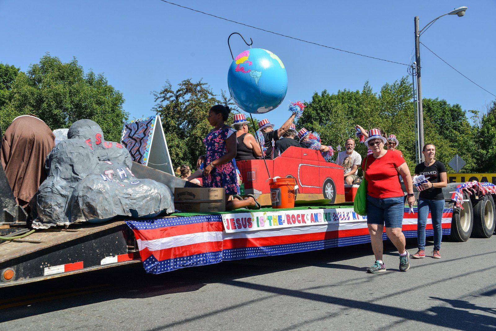 Parade float with globe, car, and people, decorated with American flags. People wearing patriotic hats. Sunny day.