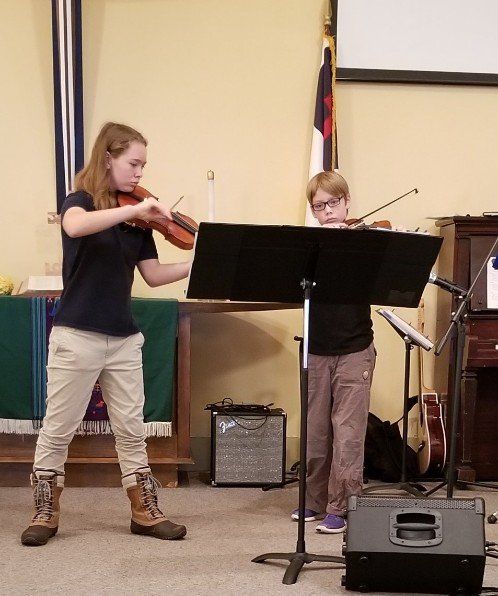 Two young people playing violins on a stage, music stand in front of them.