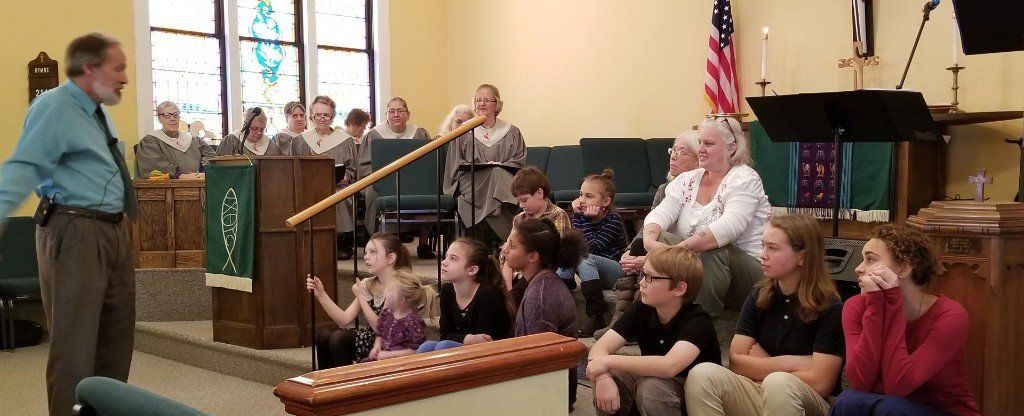 A man uses a long stick to point while speaking to a group of people in a church.