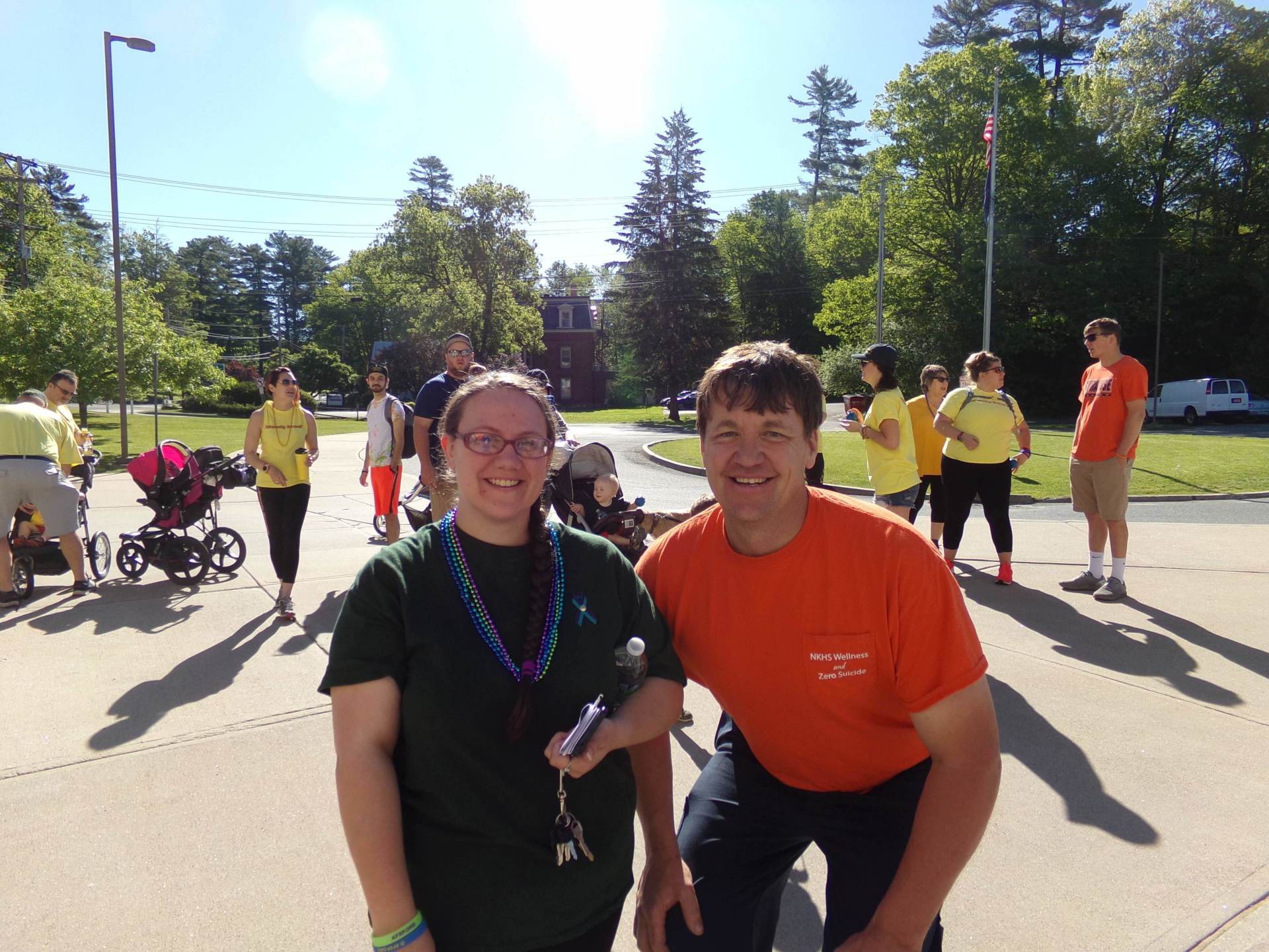 Two people in front of a group of people, possibly at an outdoor event. Green and orange shirts, sunny day.