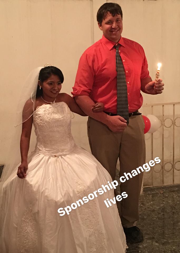 Bride in white gown and tall man in red shirt, holding a lit candle, arm in arm, smiling.