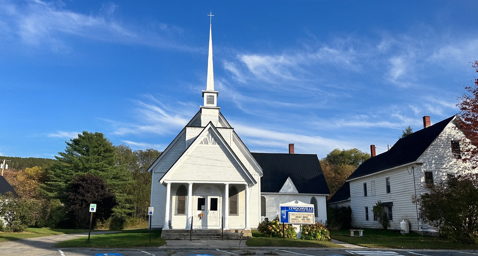 White church with tall steeple against a blue sky, tree in the foreground.