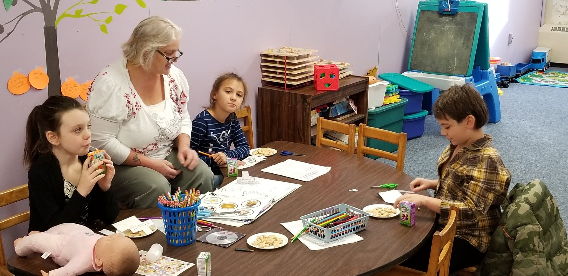 People at a table doing crafts in a room with a mural and toys.