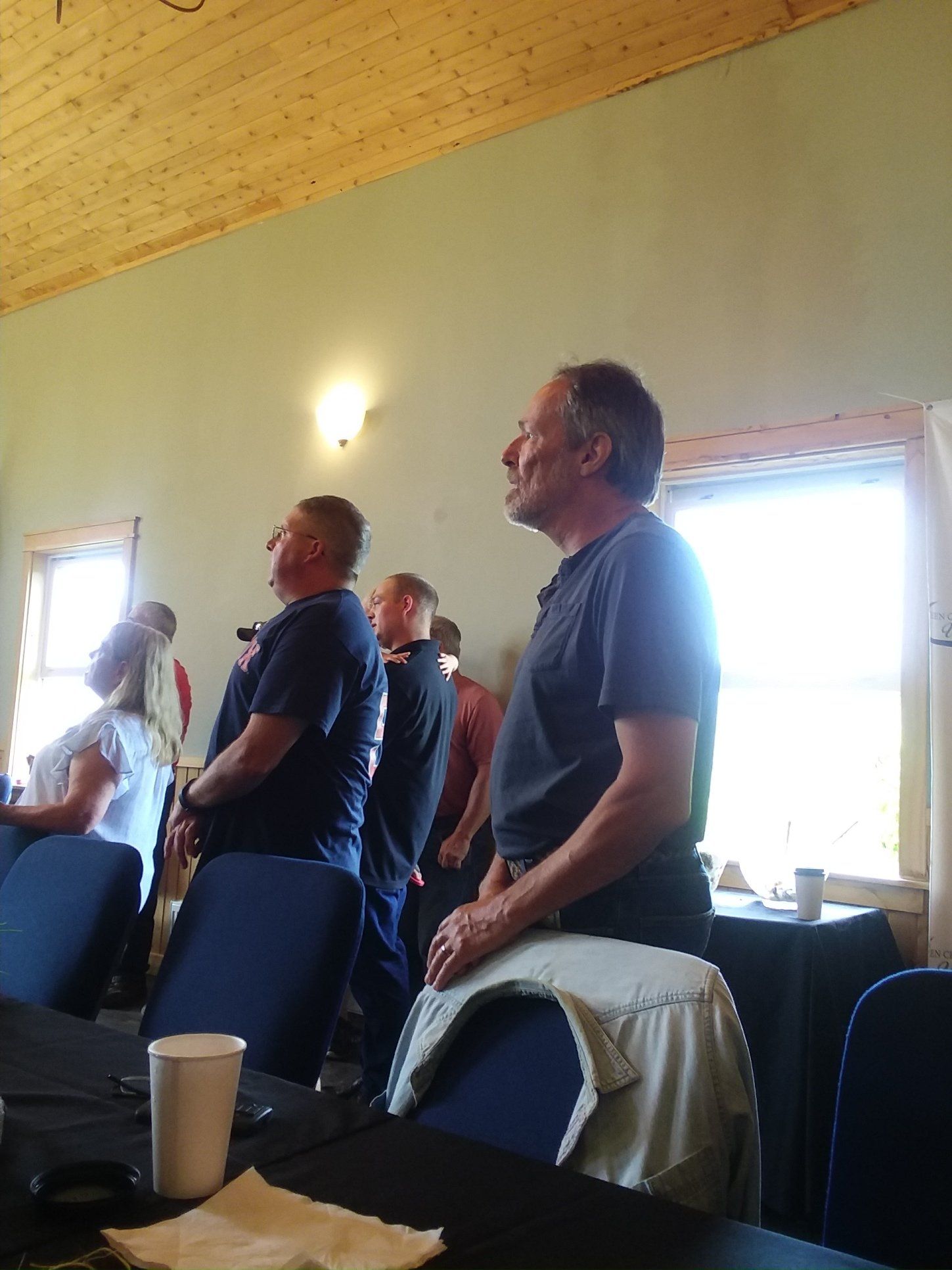 People standing, possibly at attention, in a room with a wooden ceiling and window.