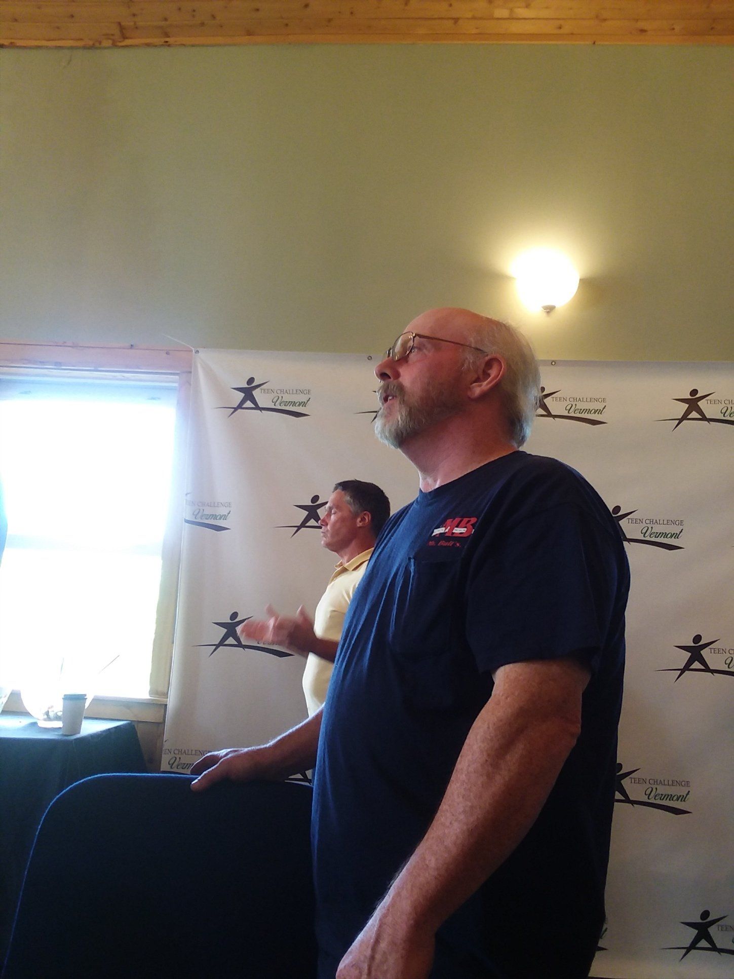 Man in blue shirt looks up, another man in yellow shirt in background. Both stand near a banner.