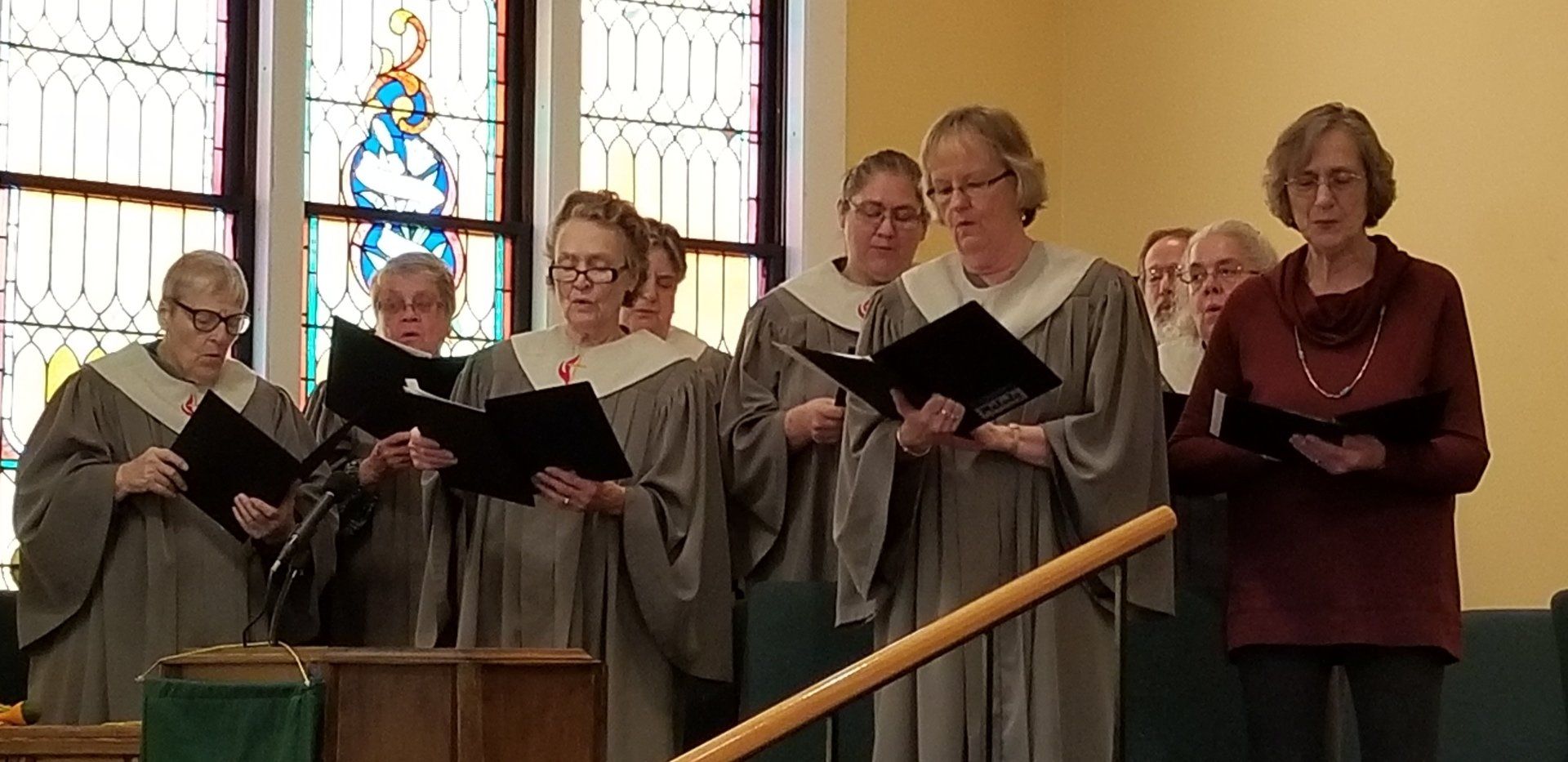 Choir in gray robes singing in front of a stained glass window. A woman in a maroon top is on the right.