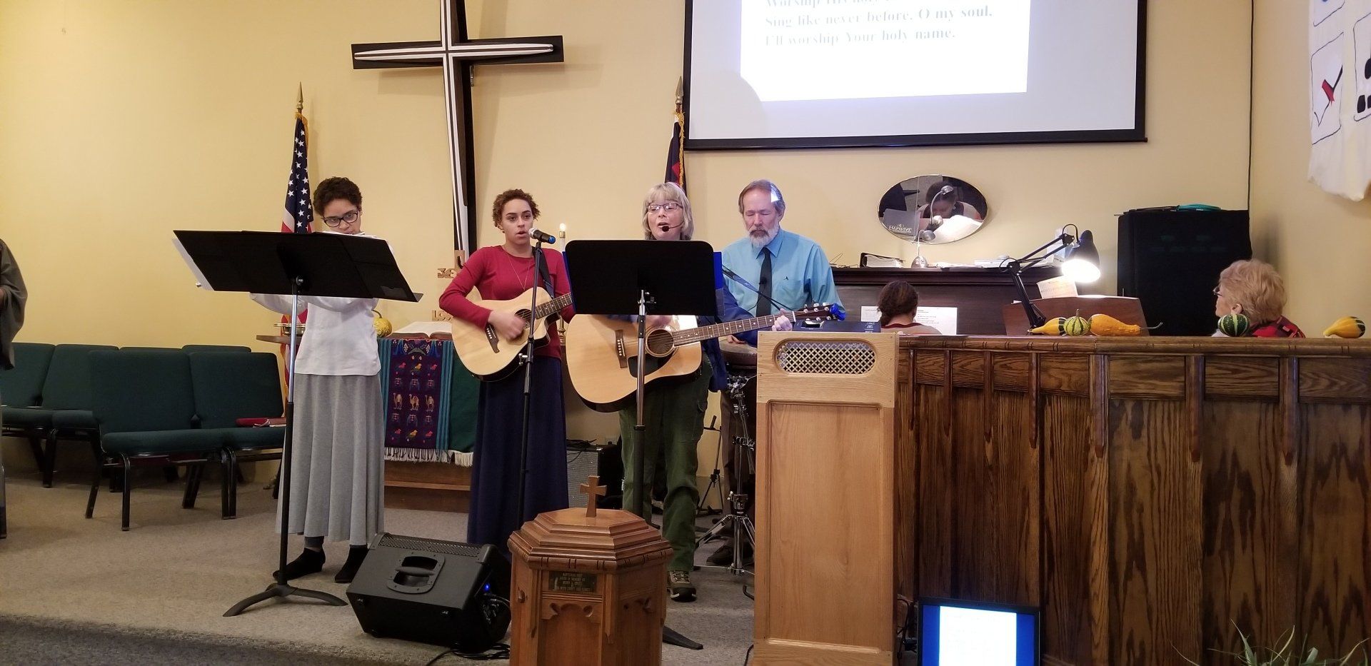 A group of people playing instruments on a stage in a church. A cross is in the background.