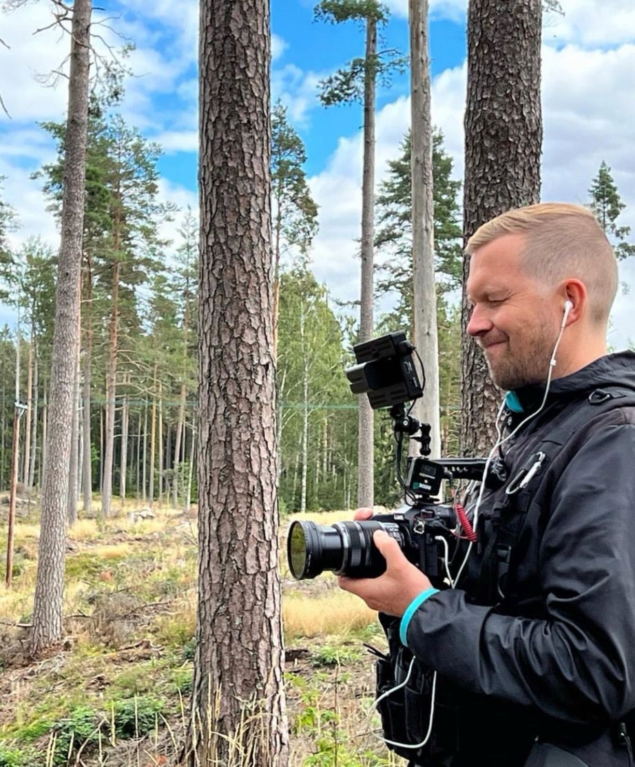 En man som filmar i en skog, håller i en kamera och bär hörlurar, med träd i bakgrunden under en blå himmel.