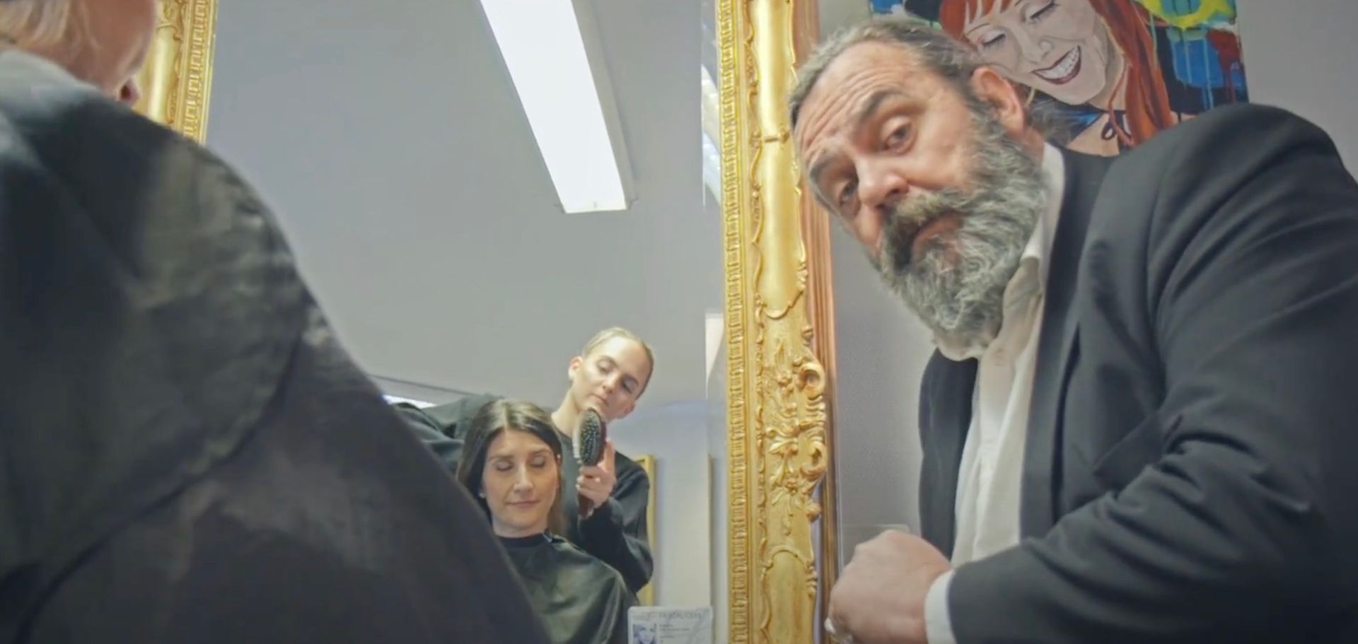 Man in suit looking into a mirror at a salon. A woman is getting a haircut, another woman is holding a comb.