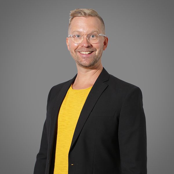 Man with glasses smiles, wearing black blazer over a yellow shirt, against a gray background.