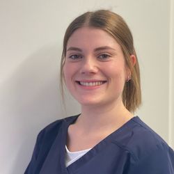 Lächelnde Frau in dunkelblauer OP-Kleidung, weißem Unterhemd, hellbraunes Haar zurückgebunden, vor einer weißen Wand.