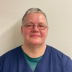 Frau mit kurzen grauen Haaren und Brille in blauer OP-Kleidung, lächelnd.