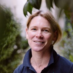 Eine Frau mit kurzen blonden Haaren lächelt in einem dunkelblauen Poloshirt in einer Gartenlandschaft mit grünem Laub.