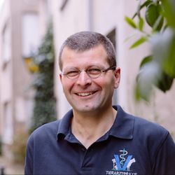 Ein Mann mit Brille lächelt und trägt ein dunkelblaues Poloshirt mit dem Logo einer Tierarztpraxis.