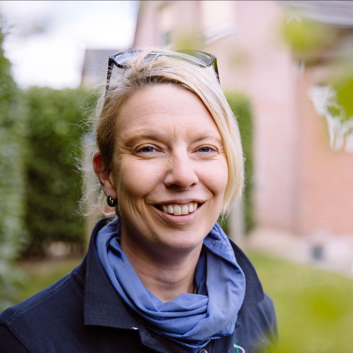 Eine Frau mit blonden Haaren lächelt. Sie trägt einen blauen Schal und eine schwarze Jacke. Sie steht draußen vor einer grünen Hecke und einem Gebäude.