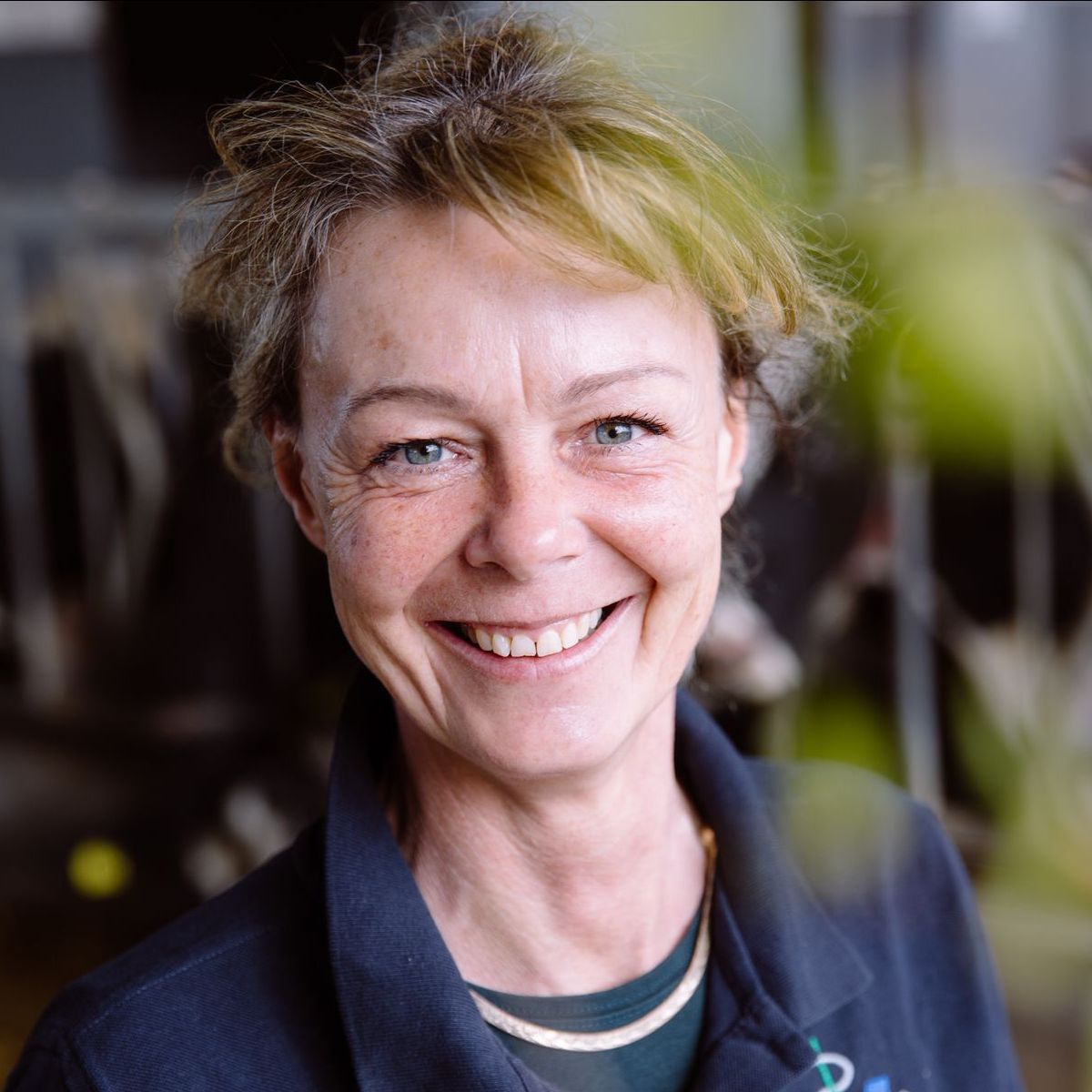 Eine Frau mit kurzen, hellbraunen Haaren lächelt in die Kamera; sie trägt eine dunkle Jacke und ein türkisfarbenes Hemd.