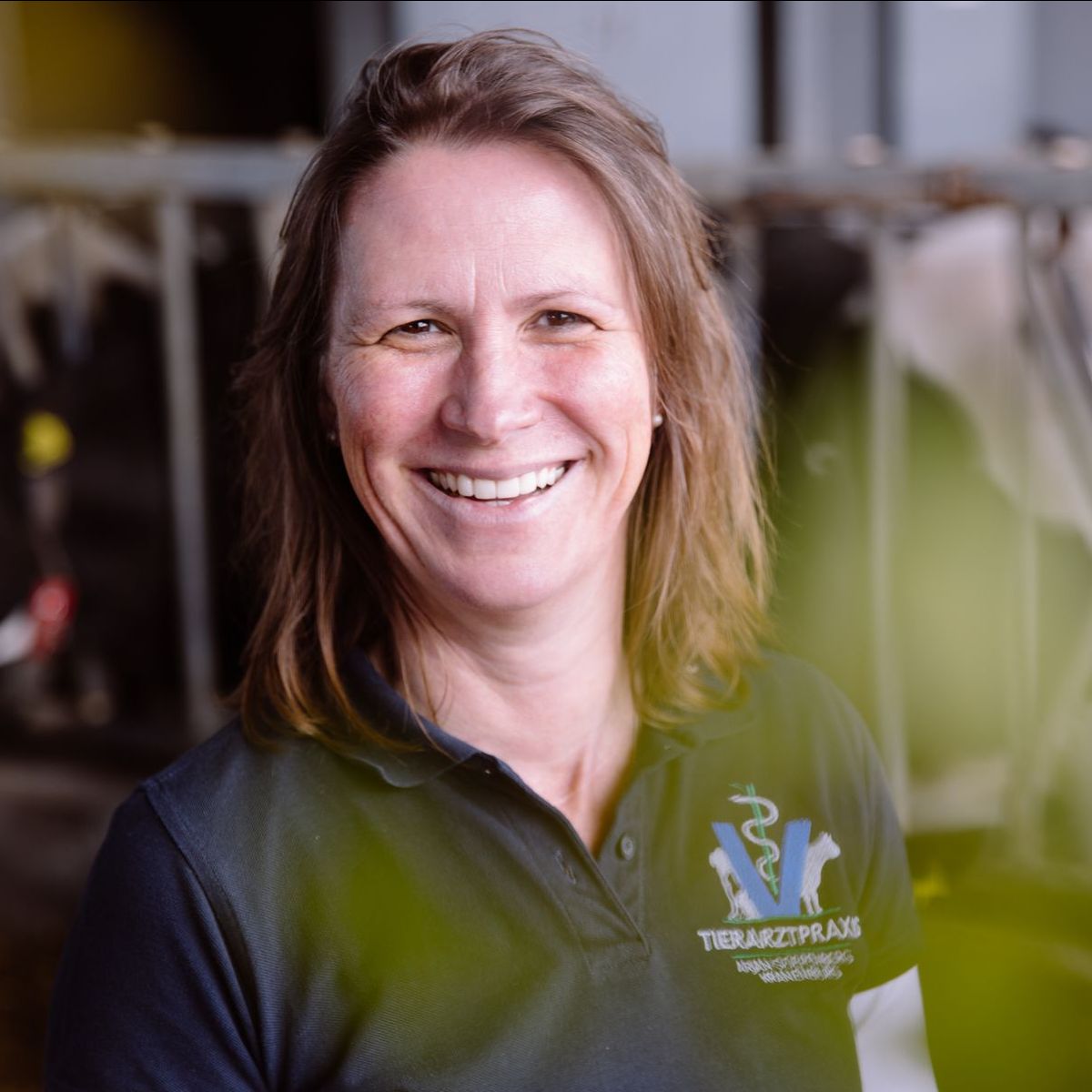 Frau lächelt, trägt ein Poloshirt mit Tierarzt-Logo und befindet sich in einem Innenraum in der Nähe von Nutztieren.