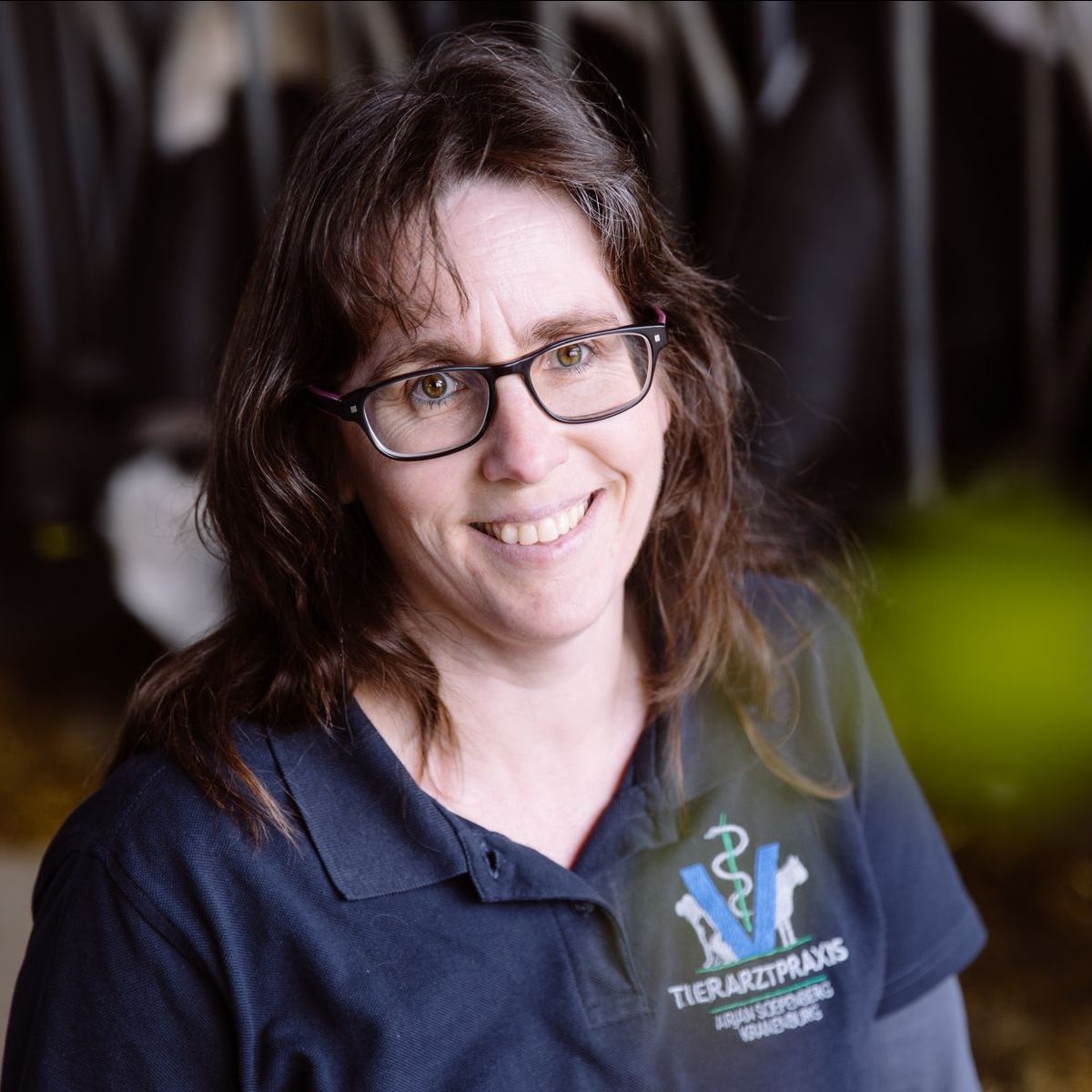 Frau mit Brille lächelt, trägt ein blaues Hemd mit einem Tierarzt-Logo, in einem Innenraum.