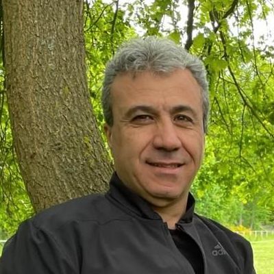 Mann mit grauem Haar, lächelnd an einen Baum gelehnt, trägt eine schwarze Jacke. Im Freien, im Hintergrund Grün.