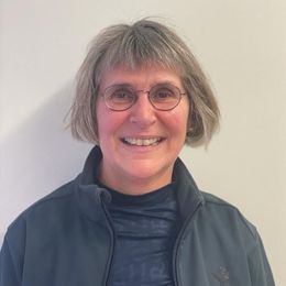 Frau mit Brille, kurzem grauen Haar, lächelnd, trägt eine dunkle Jacke, vor einer weißen Wand.