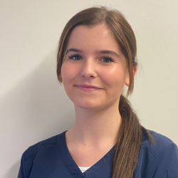 Frau in blauer OP-Kleidung lächelt, die Haare zu einem tiefen Pferdeschwanz gebunden, vor einer weißen Wand.