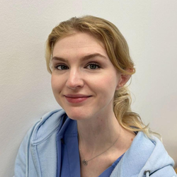 Frau mit blonden Haaren, lächelnd, trägt einen hellblauen Kapuzenpullover, vor einer weißen Wand.