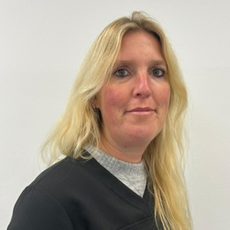 Frau mit blonden Haaren, die ein graues Oberteil und eine schwarze Jacke trägt, lächelt vor einem weißen Hintergrund.