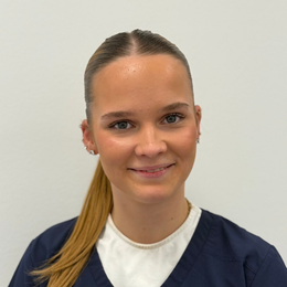 Frau mit blondem Pferdeschwanz, in blauer OP-Kleidung, lächelt.