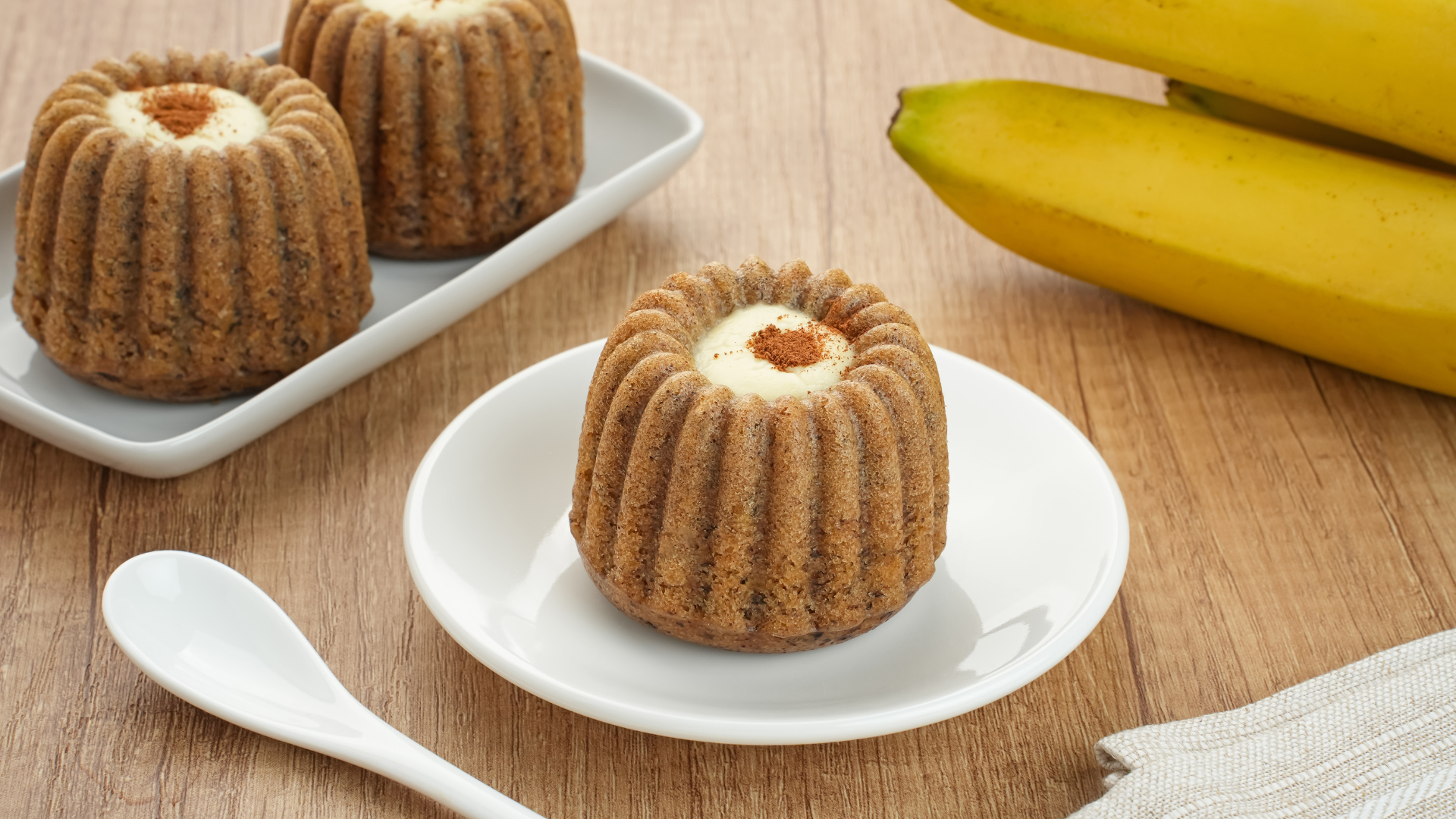 Mini bundt cakes on a white plate with banana and spoon on a wooden table.