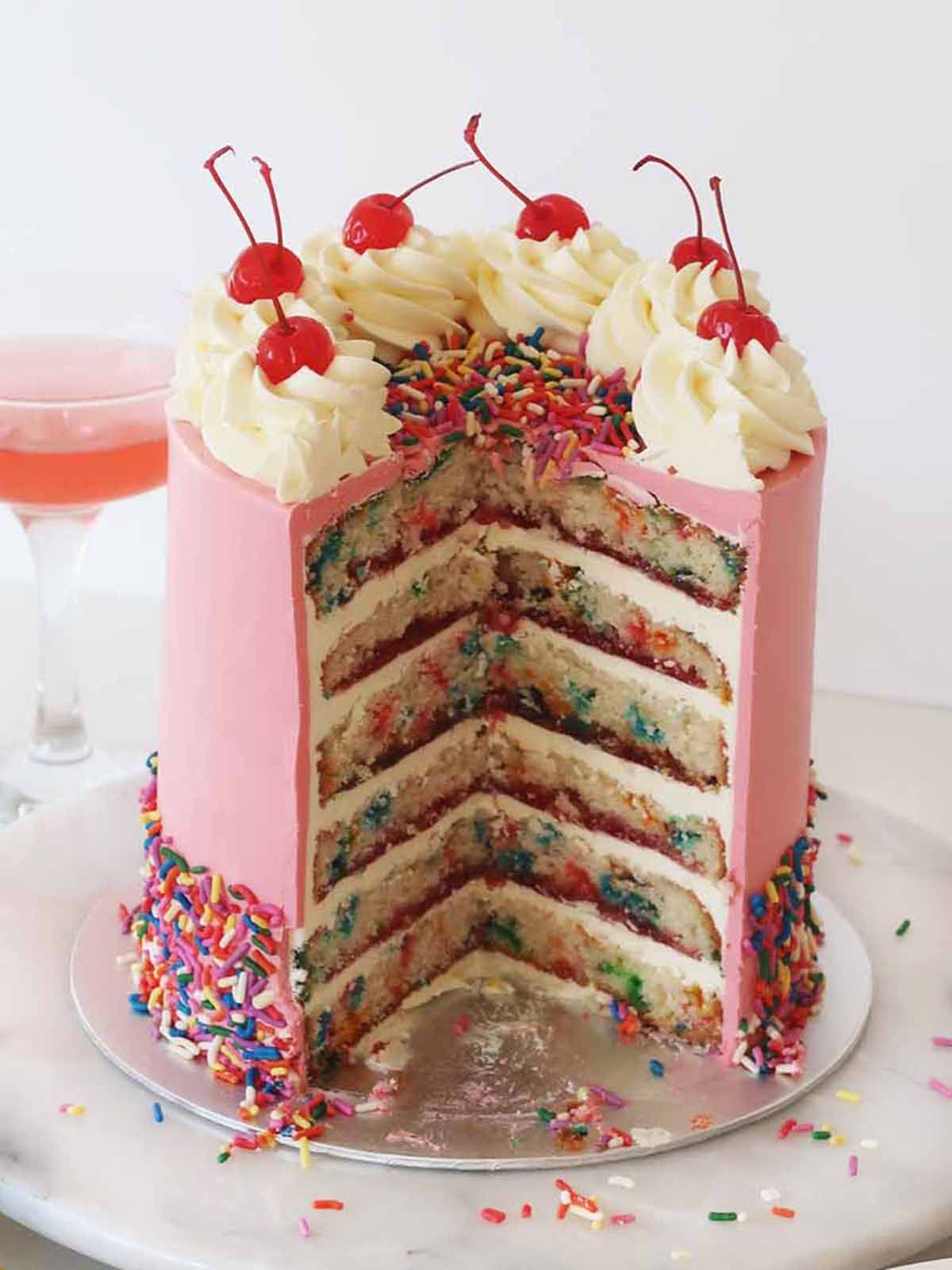 Pink frosted birthday cake with layers, sprinkles, whipped cream, and cherries.
