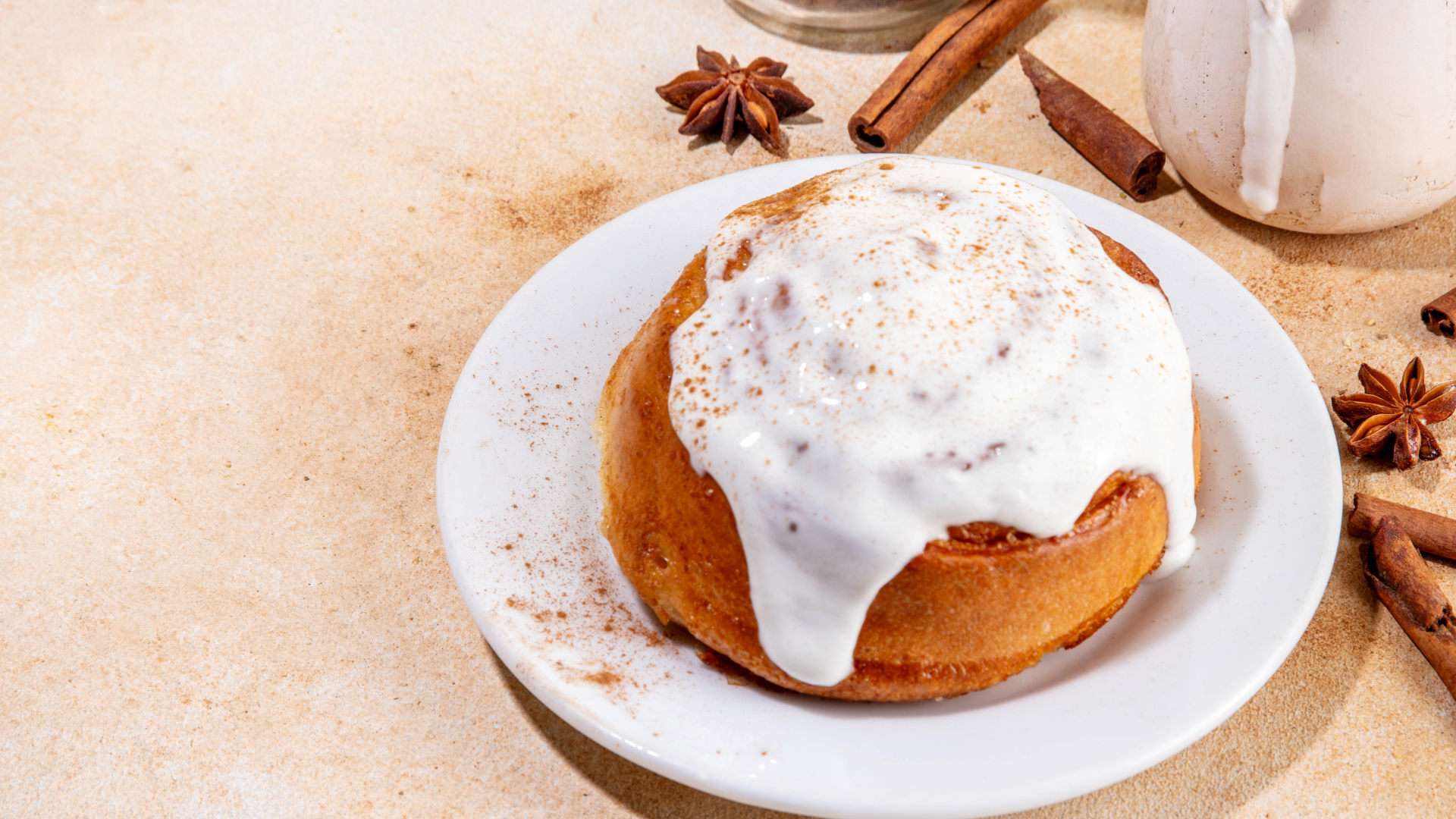 Cinnamon roll on a white plate, frosted with white icing, surrounded by cinnamon sticks and star anise.